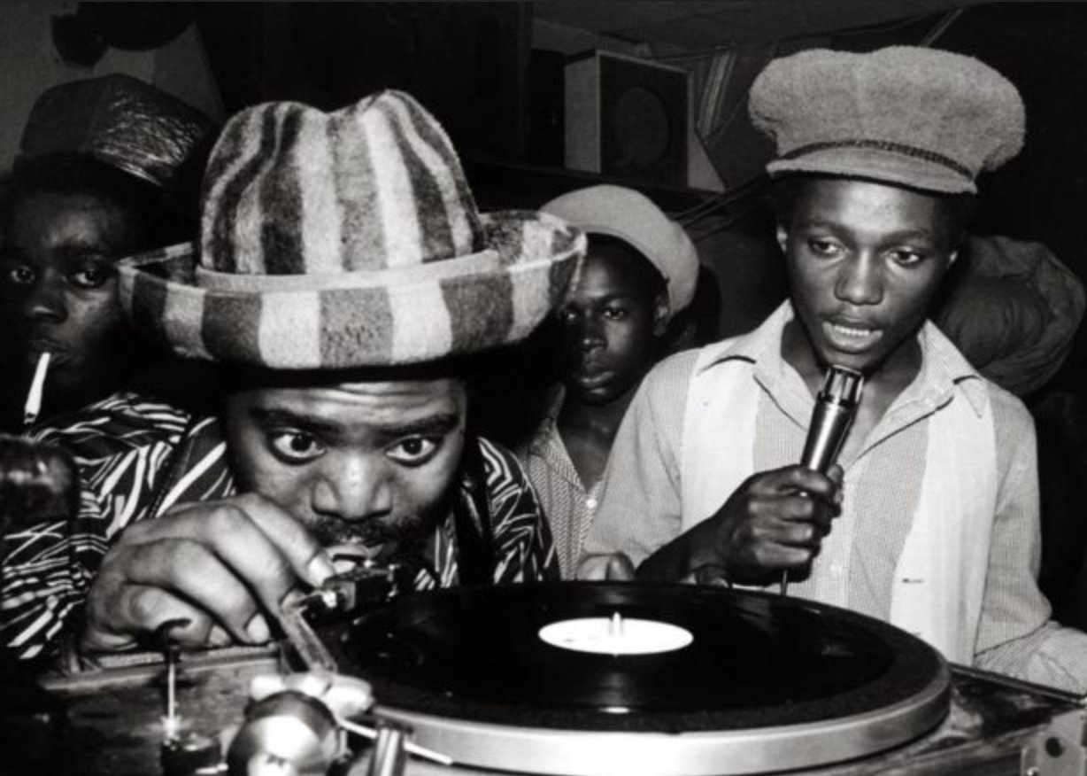 A black man in a striped hat looks closely at a record player while he adjusts the needle.