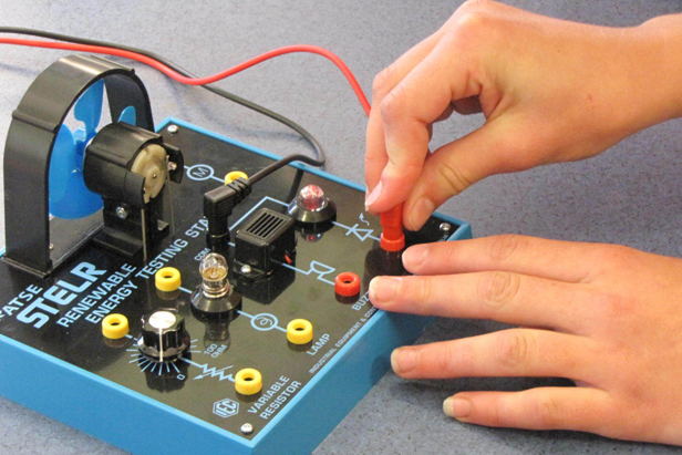 Two hands of a child plugging an electrical lead into a mini testing station featuring light bulbs, a fan and a buzzer.