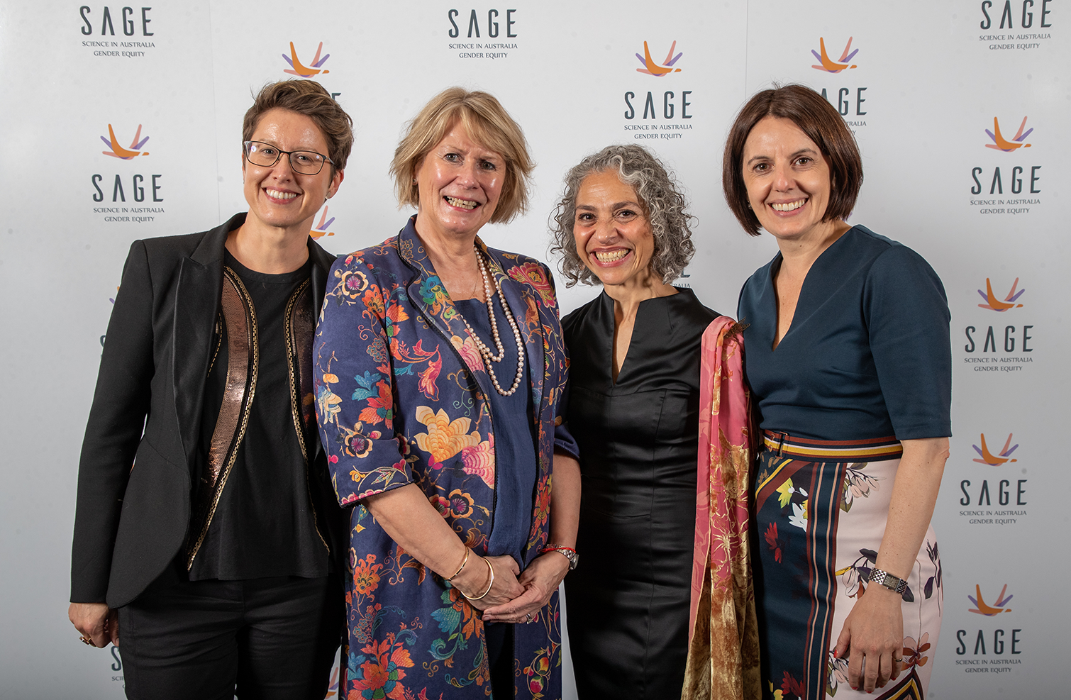 Four women standing together in fancy outfits with big smiles at the camera. Behind them is a wall with the SAGE logo.