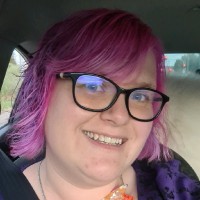 Sarah Crowe has pink and purple hair and wears black glasses. She is looking at the camera and smiling.