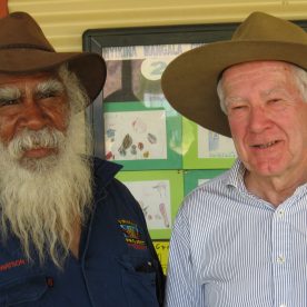 An Aboriginal man with a white beard on the left stands next to a white man on the right, both are wearing hats.